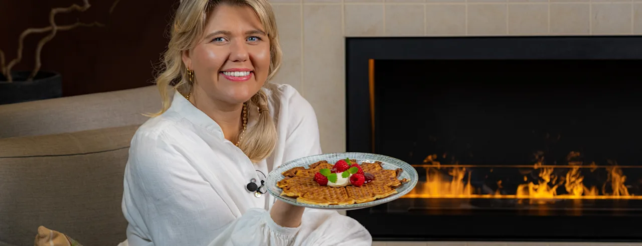 Våfflor med bär och grädde på ett mönstrat bord, med en hand som håller en kopp kaffe.