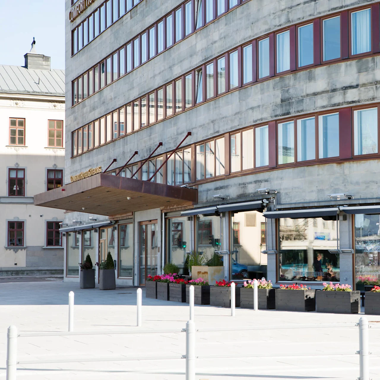 Byggnadsfasad visar flera fönster och skyltar med texten "COMFORT HOTEL GÖTEBORG" och "BAR & RESTAURANT." Blomsterlådor pryder ingångsområdet i en stads miljö.