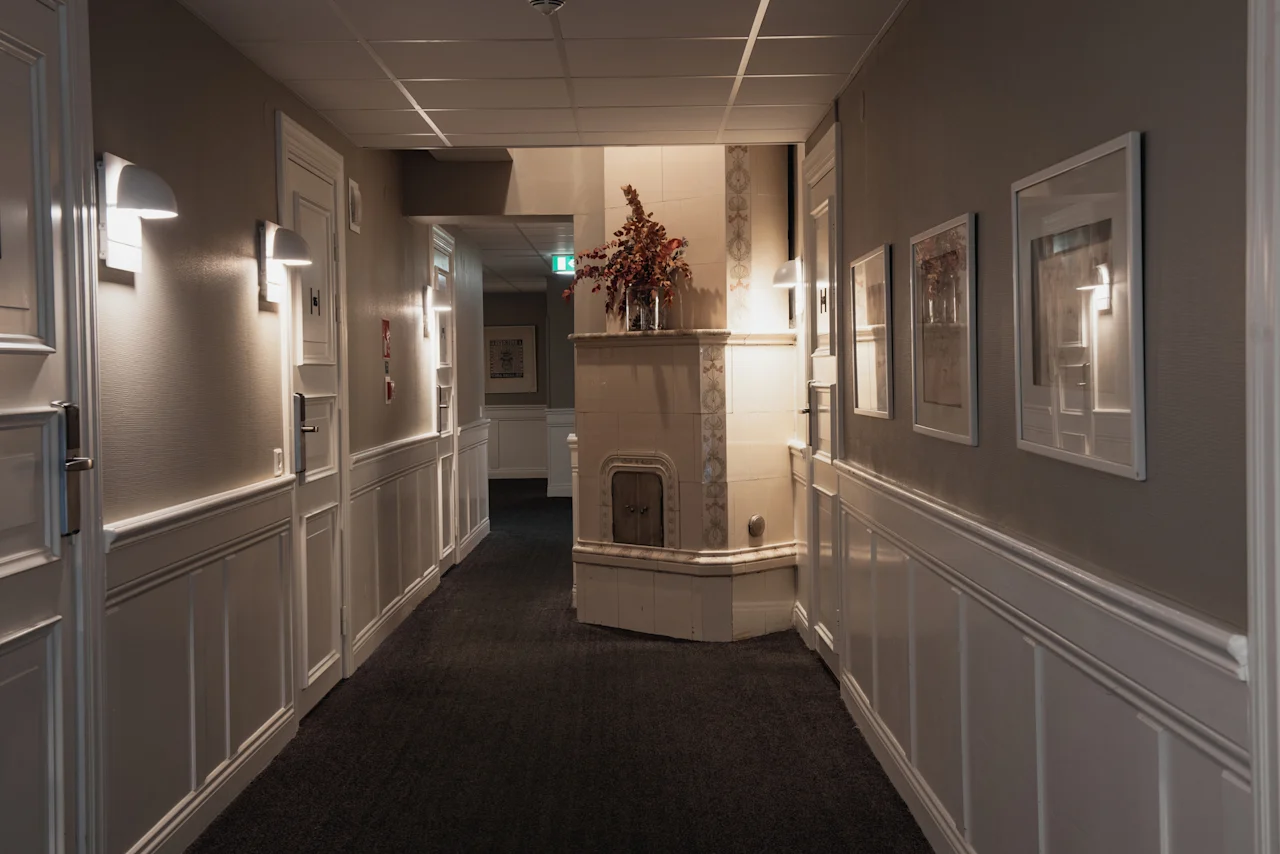 En väl upplyst hotellkorridor med dörrar på båda sidor, med en dekorativ öppen spis och inramad konst på väggarna.