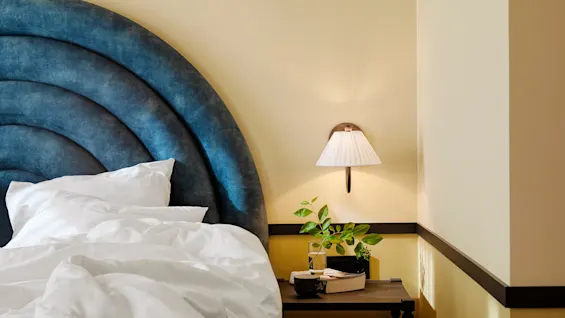 A bed with a blue tufted headboard and white sheets is beside a nightstand with plants and books under a wall-mounted lamp in a softly lit room.
