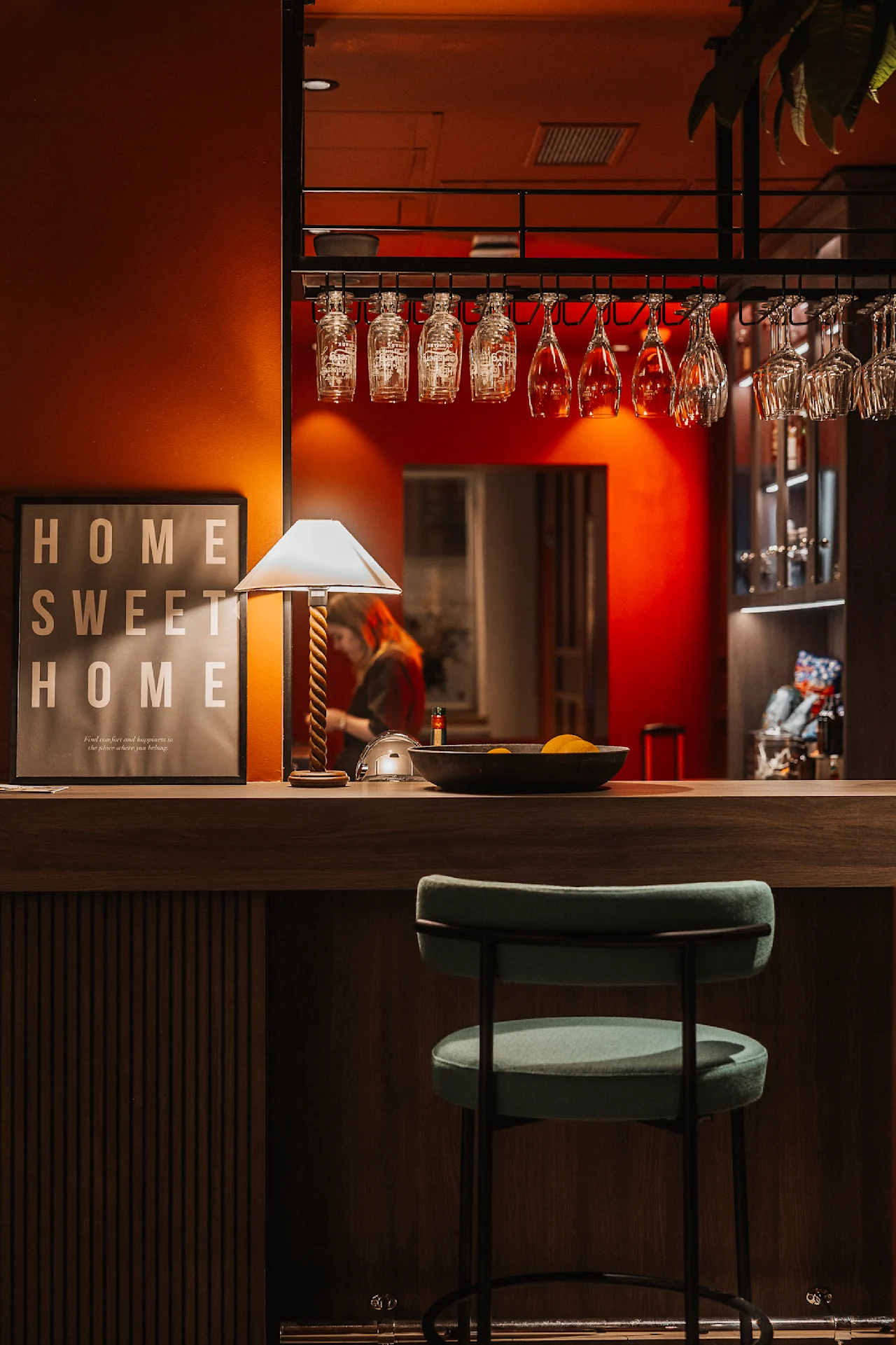 En køkkenbar med en grøn stol, belyst af en lampe, har hængende glas og et "HOME SWEET HOME" skilt. En person ses i baggrunden.