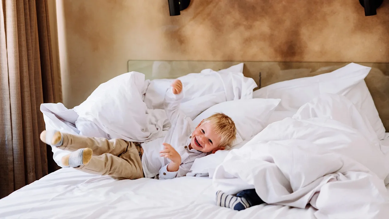 A young child is joyfully playing and rolling on a bed covered with white pillows and blankets in a cozy, warmly lit room.