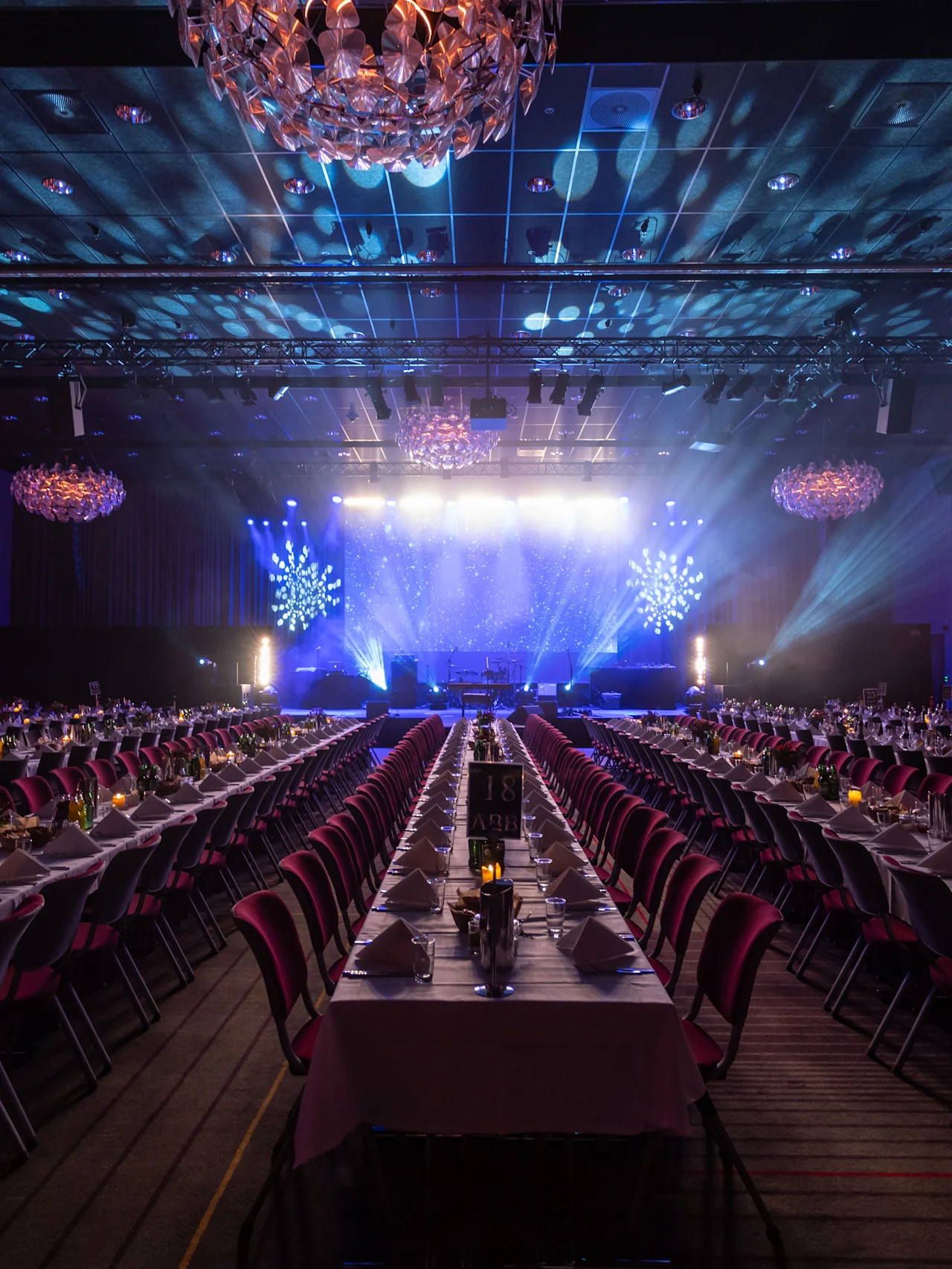 Tables with white tablecloths are neatly arranged in long rows, facing a brightly lit stage with blue lighting and decorative snowflakes. The setting is an elegant banquet hall.