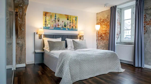 A neatly made bed with white bedding is in the center of a room featuring exposed brick walls and a painting above the headboard, illuminated by wall lamps and natural light from a window.