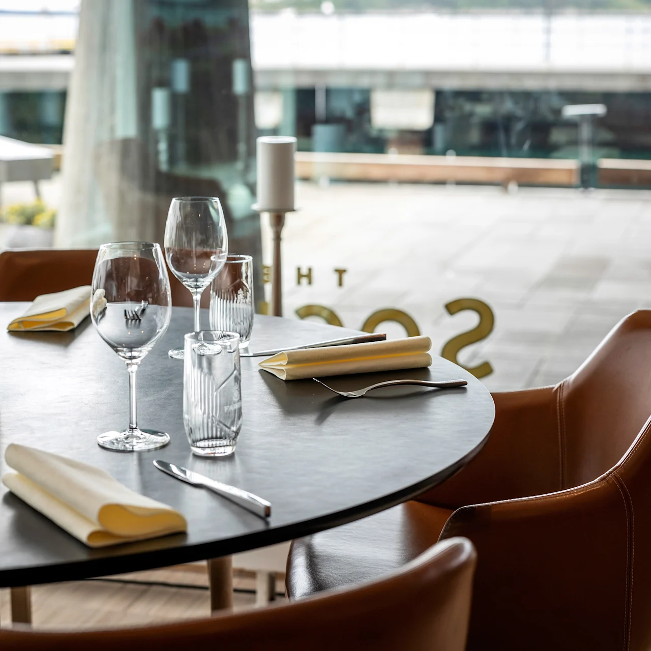 Rundt bord dukat med prydligt ordnade glas, bestick och servetter; omgivet av bruna stolar i en modern restaurangmiljö med stora fönster. Text på glas: "THE SSOS" (delvis synlig).