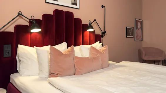 Et stilfullt soverom med en plysjseng med rød fløyels hodegavl, mykt rosa og hvitt sengetøy, og varm belysning fra vegglamper.
