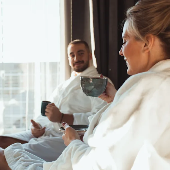 Two people in bathrobes relaxing by a window, enjoying drinks. One person has their feet up on the bed, looking comfortable.