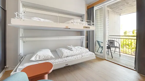 A modern bunk bed setup in a bright room, with the lower bed made and the top bunk folded against the wall, near an open balcony door.