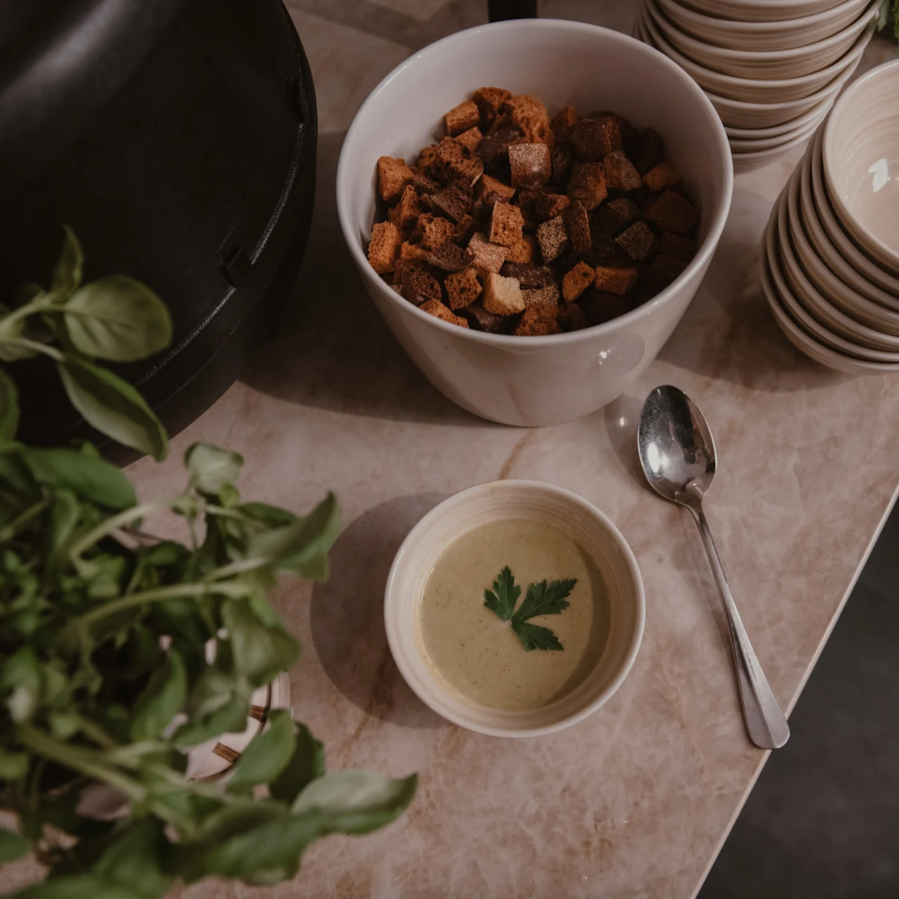 A small bowl of soup with a garnish sits on a kitchen counter beside a spoon, croutons, stacked empty bowls, and fresh basil.