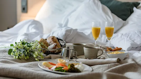 Breakfast tray holds croissants, mugs, plates of cheese and salad, glasses of orange juice, and flowers. Set on a bed with white linens.