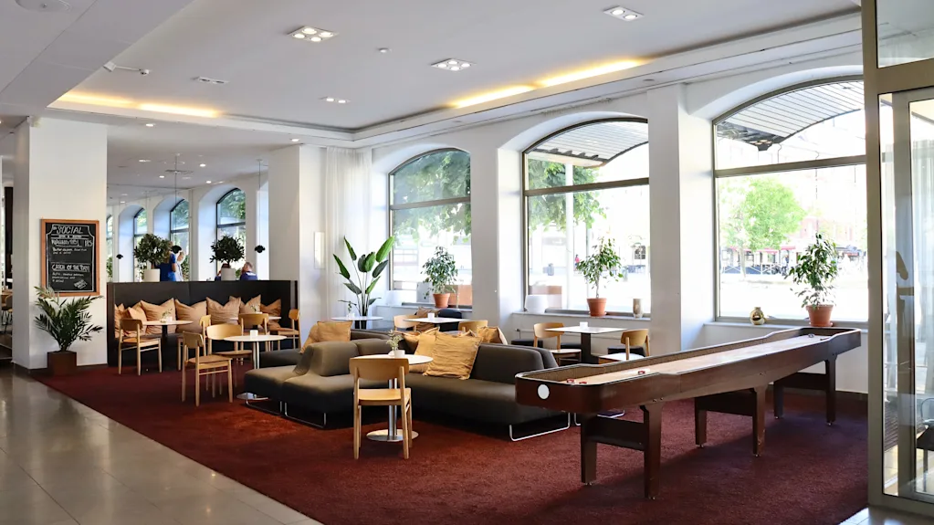 Norsk bokmål: Loungeområde med sofaer og stoler på et rødt teppe, med planter og store vinduer mot gaten. Et shuffleboardbord kompletterer det moderne, koselige interiøret.