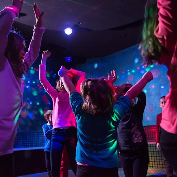 Børn der danser med hænderne i vejret under farverige blinkende lys til et minidisco.