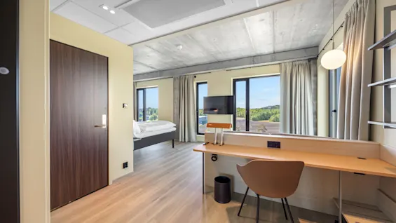 A modern hotel room with a bed, a desk, a TV, and large windows offering a scenic view.