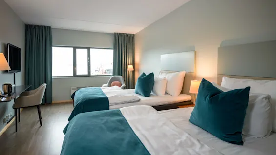 To senge med turkis detaljer er pænt redt ved siden af hinanden i et hotelværelse med vindue, skrivebord med stol og lamper til belysning.