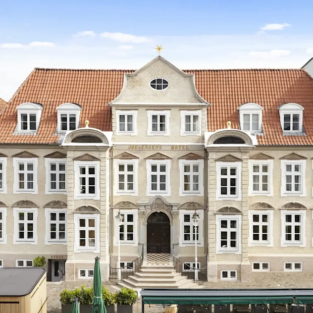 En storslået, historisk hotelbygning med rødt tegltag, mange vinduer og en fremtrædende indgang. Teksten HOTEL er synlig.