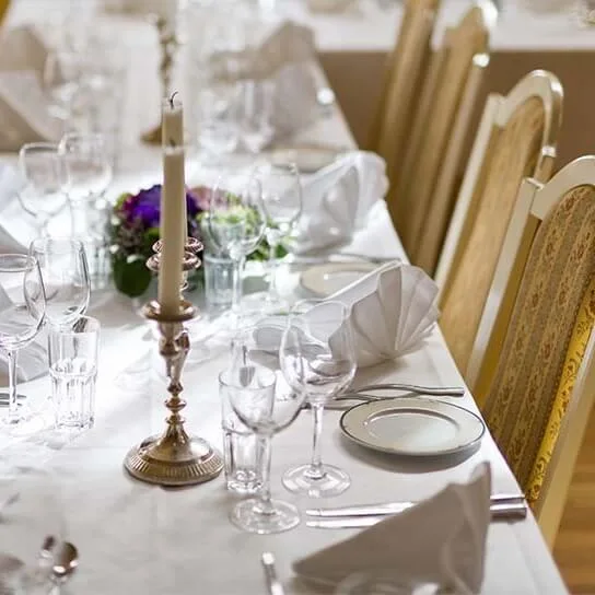 Et elegant dækket spisebord viser foldede hvide servietter, tomme glas og hvide tallerkener, omgivet af træstole. En sølvlysestage og lilla blomster tilføjer dekorative elementer.