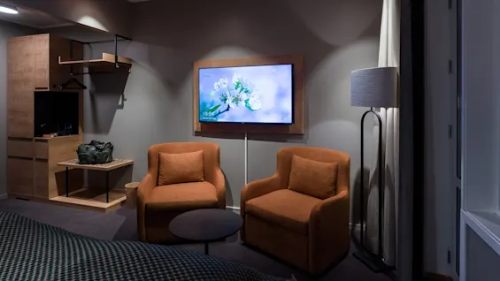 A pair of orange armchairs face a wall-mounted TV displaying white flowers and text: "Philips Professional Display, 16:54, Wednesday, 22 December." Nearby, a wooden shelf and a standing lamp.  