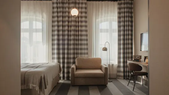 A cozy hotel room with a bed, armchair, desk, and large windows with striped curtains. Exposed wooden beams on the ceiling.