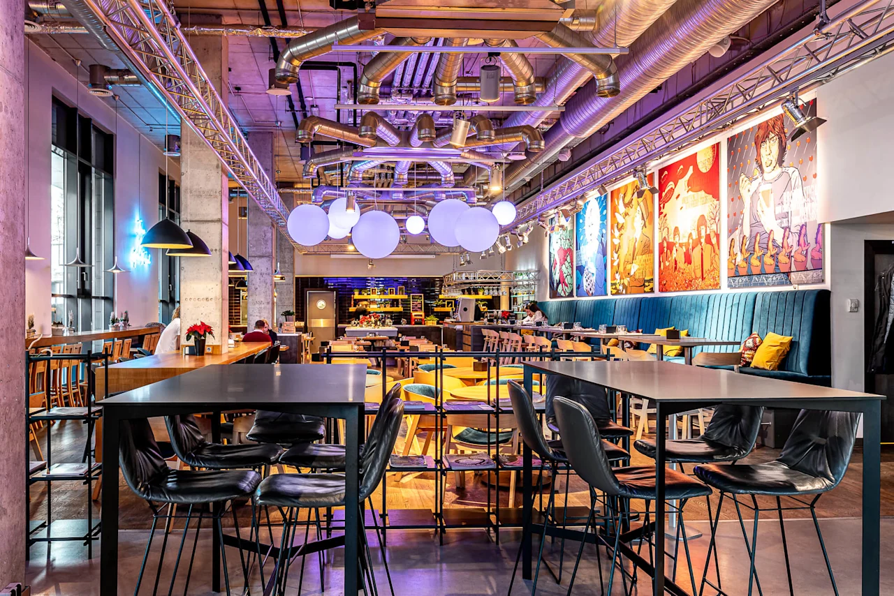 Modern dining space with chairs and tables, vibrant artwork on the walls, and large, spherical lights overhead. Industrial decor with exposed ducts and colorful ambiance.