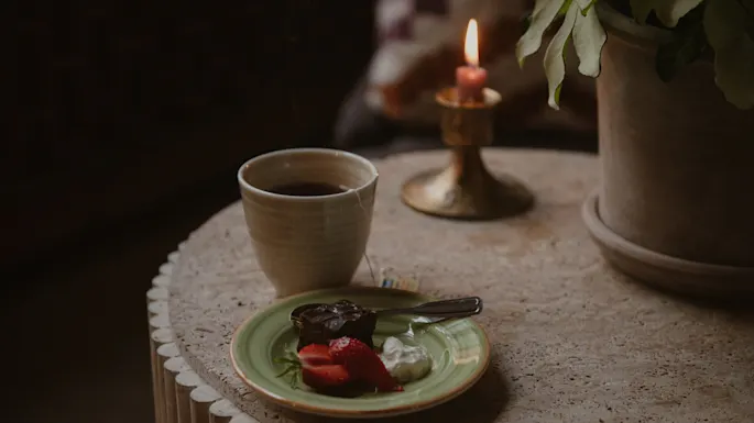En kopp te på et rundt bord med en tallerken som inneholder kake, jordbær og krem. Et tent lys og en plante i bakgrunnen skaper en koselig atmosfære.