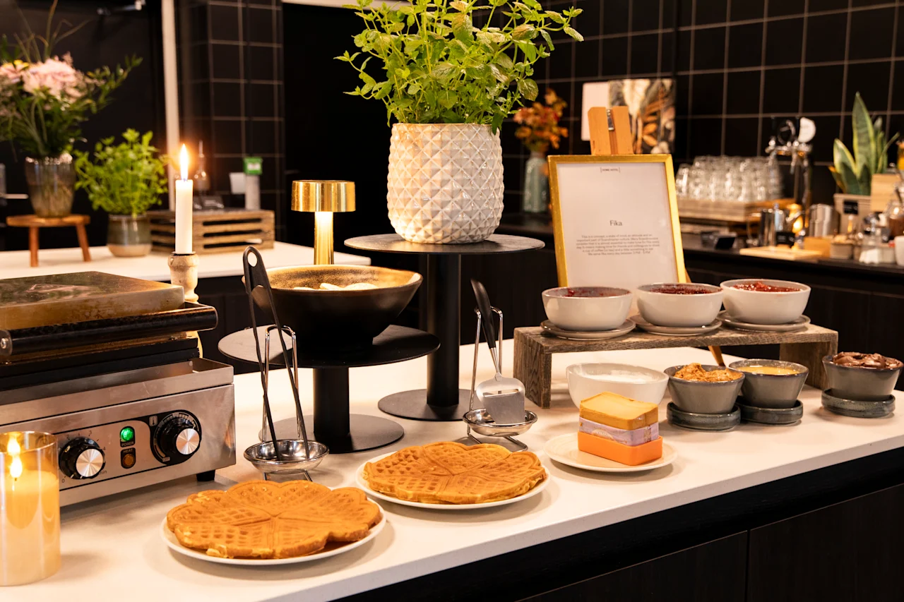 En koselig vaffelstasjon med vaffeljern, nystekte vafler og diverse tilbehør som syltetøy og krem. Et skilt sier Fika.