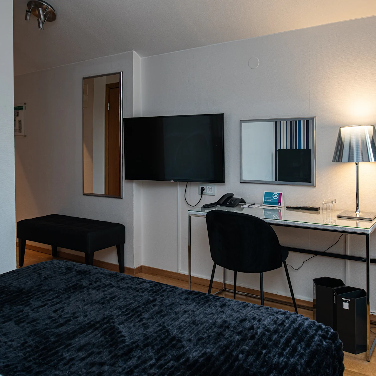 Et ryddeligt hotelværelse har et skrivebord med stol, en tændt lampe og et vægmonteret TV. En seng med mørkt sengetæppe er i forgrunden.