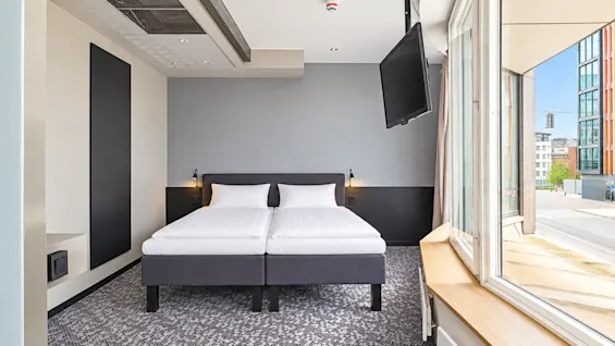 Bright hotel room featuring a double bed, wall-mounted TV, and large windows overlooking the street.