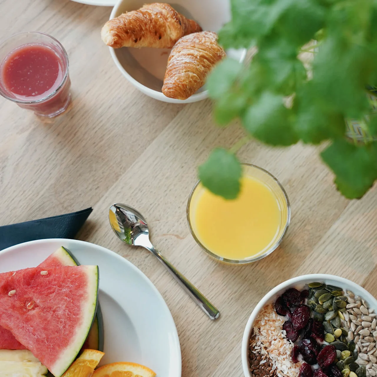 En frokost på et trebord inkluderer croissanter, vannmelonskiver, en blandet bolle med frø og bær, glass med appelsin- og rød juice, og grønne planter.
