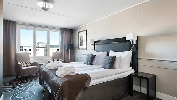 A neatly made bed in a well-lit hotel room with modern decor, two armchairs, and a city view through the windows.