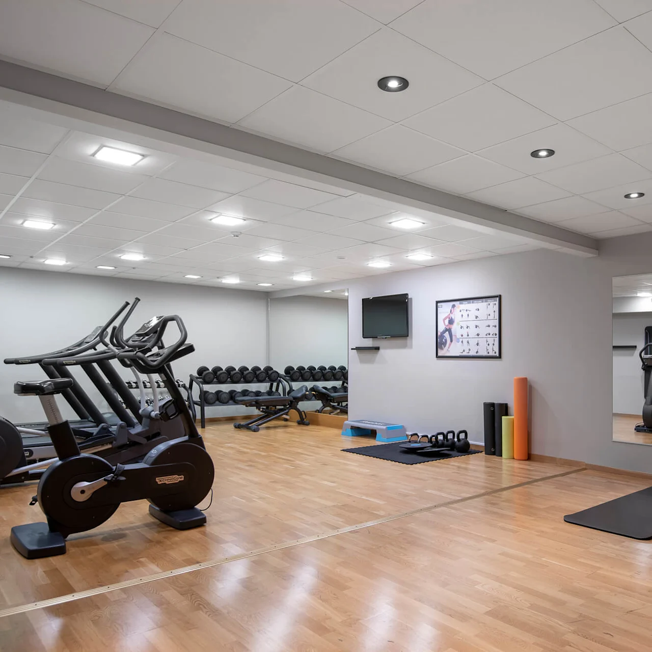 Löpband står stilla i ett ljust gym, omgivet av vikter, mattor och en spegel som speglar utrustningen. På väggarna finns affischer och en TV, med trägolv under fötterna.