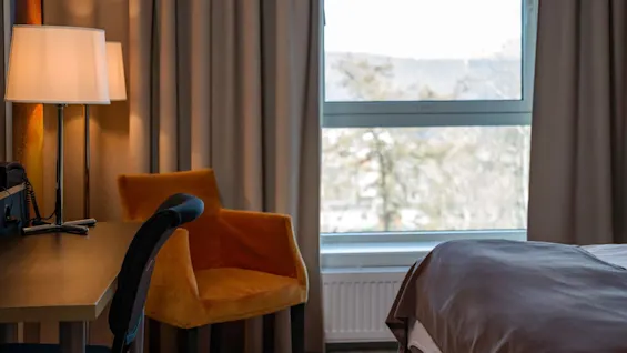 En skrivebordslampe oplyser et bord ved siden af en sort stol og en orange lænestol. Et stort vindue med gardiner viser en sløret udendørsscene.