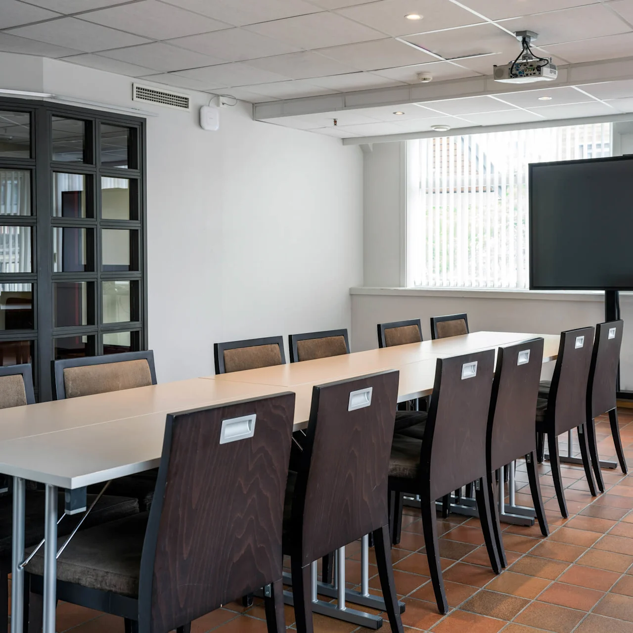 A long conference table with brown chairs surrounds a modern meeting room. On the wall, a flat-screen TV stands in front of large windows with blinds.