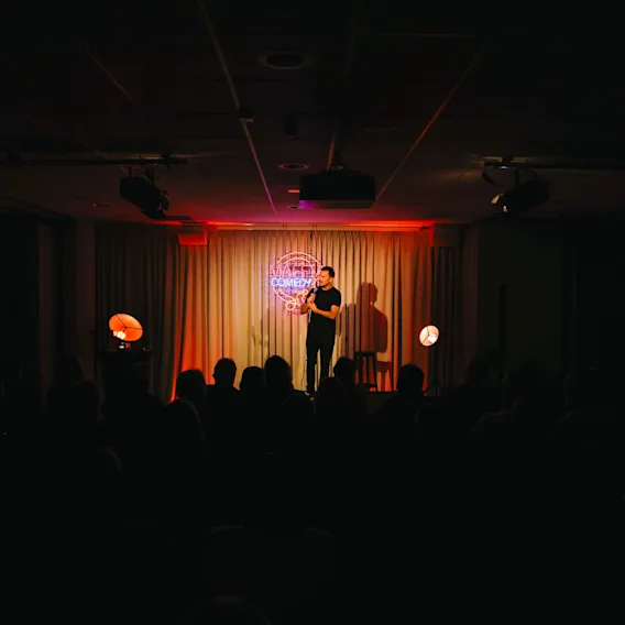 A stand up comedian performing on a dimly lit stage in front of a seated audience.