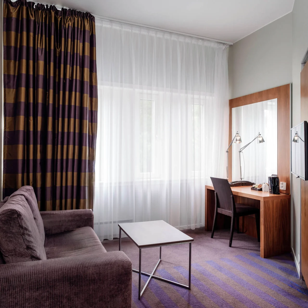 En lilla sofa vender mod et lille bord i et hotelværelse med stribede gardiner. Et skrivebord holder to lamper og en stol ved vinduet. Ingen synlig tekst.
