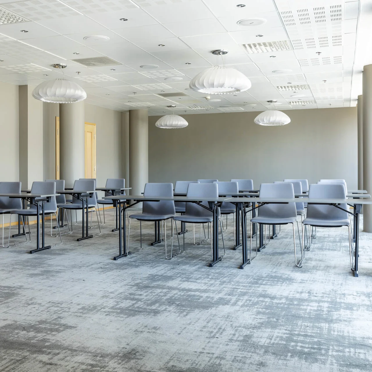 Rows of empty gray chairs and tables, arranged classroom-style, sit on a carpeted floor in a well-lit room with gray walls and ceiling lights.