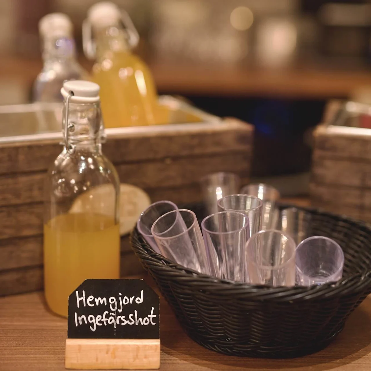 Clear glass bottles filled with yellow liquid, next to a black basket of plastic cups, labeled "Hemgjord Ingefärsshot" on a sign, in a cozy kitchen setting.