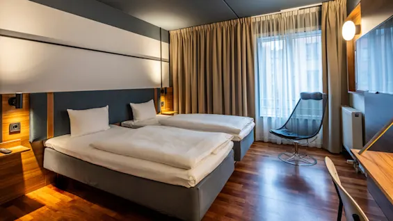 A hotel room with two single beds covered with white bedding, separated by a bedside table with a reading lamp. The room features large windows with beige curtains, a sleek chair, and a wooden desk with a minimalist chair.
