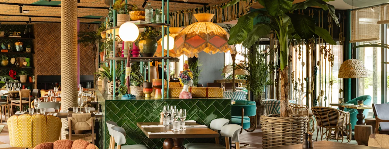 Et restaurantinteriør har fargerik, eklektisk innredning med frodige planter, tremøbler og myk belysning. Bord er dekket med glass og bestikk, og skaper en innbydende spiseatmosfære.