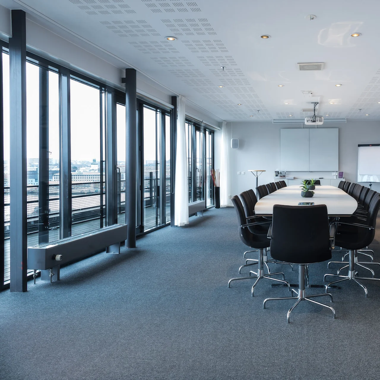A long conference table with black chairs stands in a modern meeting room. Large windows line the left side, and abstract paintings hang on white walls.
