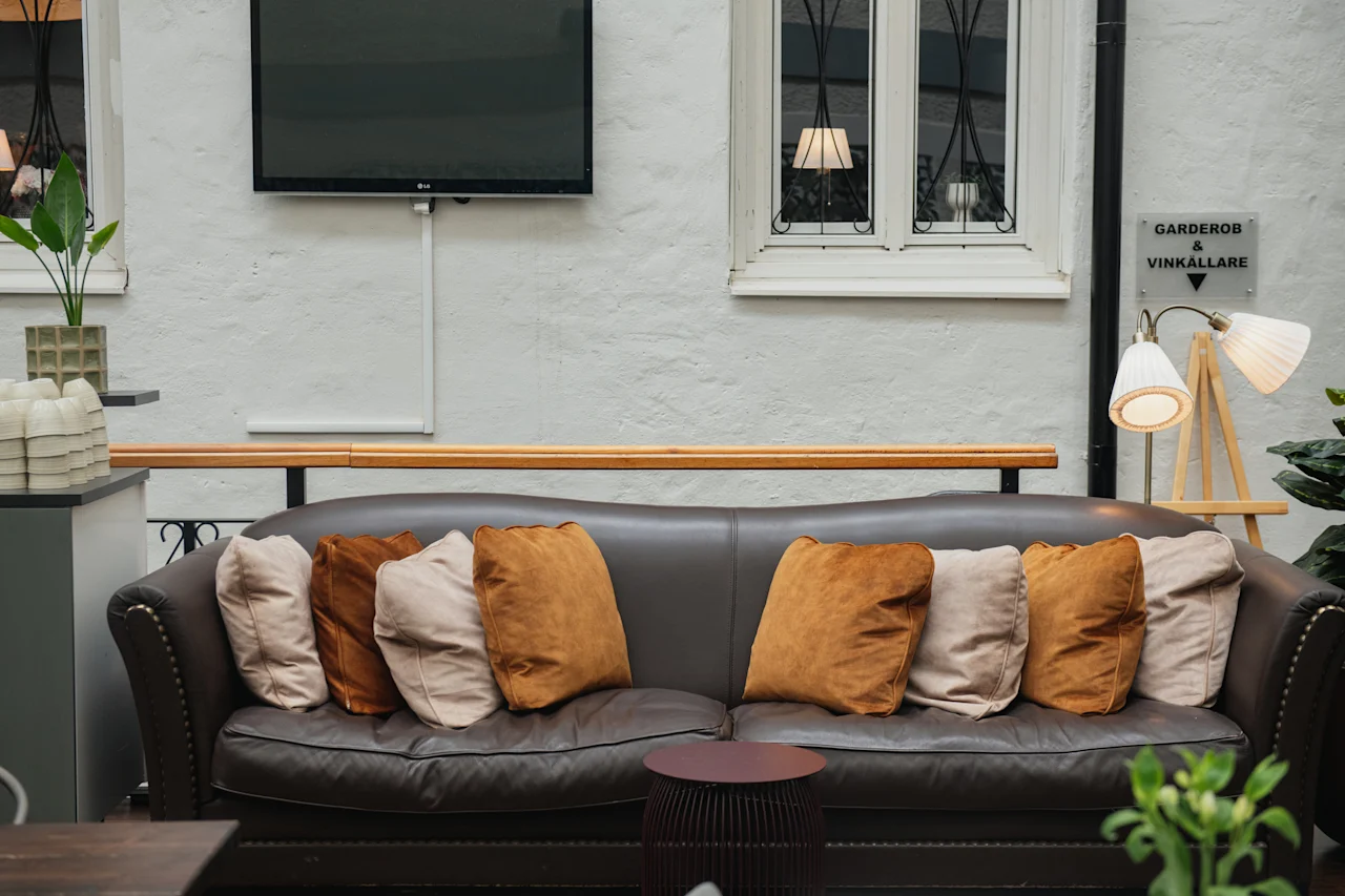 En brun lädersoffa med beige och bruna kuddar står framför en texturerad vit vägg. I närheten finns en skylt som säger "Garderobe & Vinkällare." Växter och en lampa skapar en mysig atmosfär.