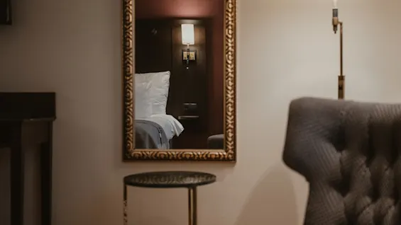 A cozy hotel room with a mirror reflecting a comfortable bed, an armchair, and a small table. add Home Hotel Kung Oscar