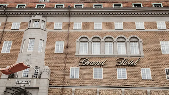 The stately brick facade of a hotel, featuring numerous windows and a prominent sign that reads 'Grand Hotel'. The entrance says 'GRAND'.