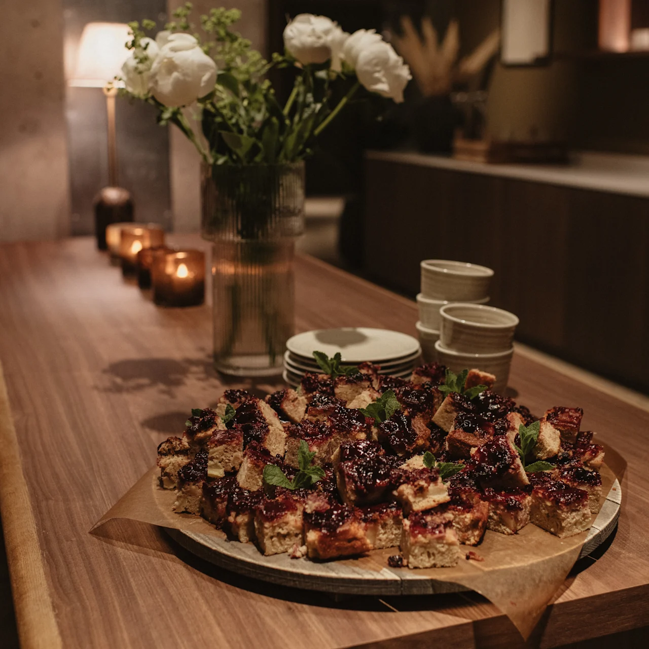Platter of bite-sized, glazed snacks garnished with herbs, placed on a wooden table in a softly lit, modern dining area; nearby, a vase of white flowers and lit candles.
