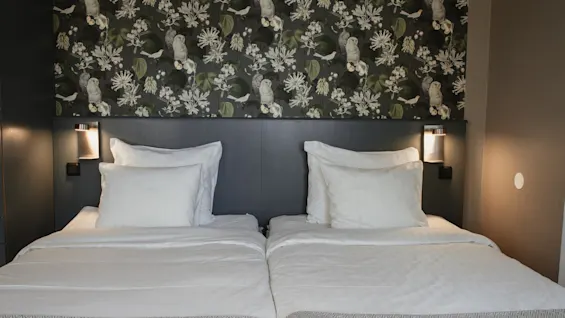 Two neatly made beds with white linens and a dark headboard, set against a patterned wallpaper with owls and leaves at Home Hotel Uppsala.
