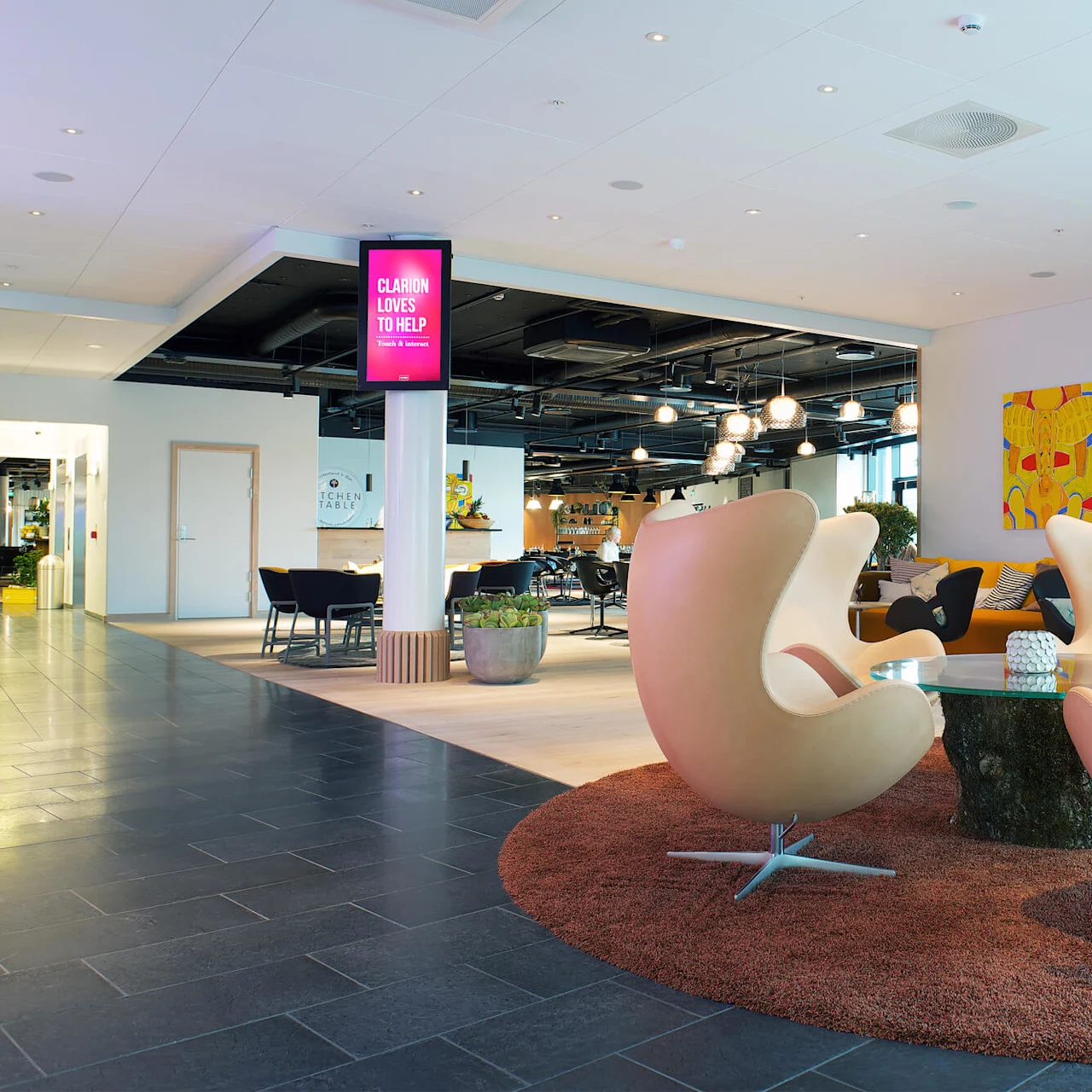 Fyra moderna stolar omger ett glasbord på en matta, i en stilren lobby med abstrakt konst. En skylt lyder: "CLARION LOVES TO HELP Travel & Business."