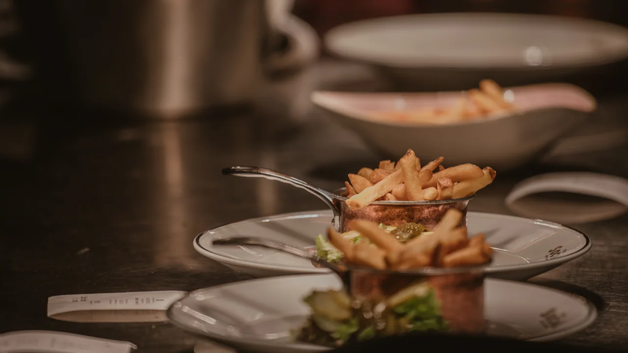 Crispy fries sit in a metal cup on a white plate, garnished with greens, in a dimly lit kitchen, surrounded by pots and other dishes. 