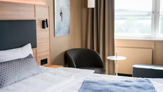 A neatly made hotel bed features a blue throw blanket, set in a modern room with a dark armchair, small round table, wall art, curtain, and window view.