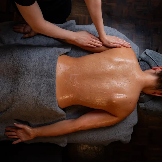 A person lies face down on a massage table, receiving a back massage with oiled skin, covered partially by a gray towel in a dimly lit room.