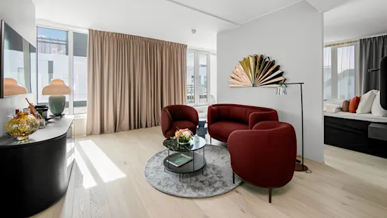 Red armchairs and a small sofa encircle a glass coffee table on a round rug in a bright room, accented by a unique wall mirror and various decorative items.