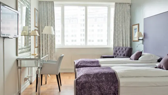 Et ryddig hotellrom med to enkeltsenger, et skrivebord og store vinduer som viser bygninger utenfor.